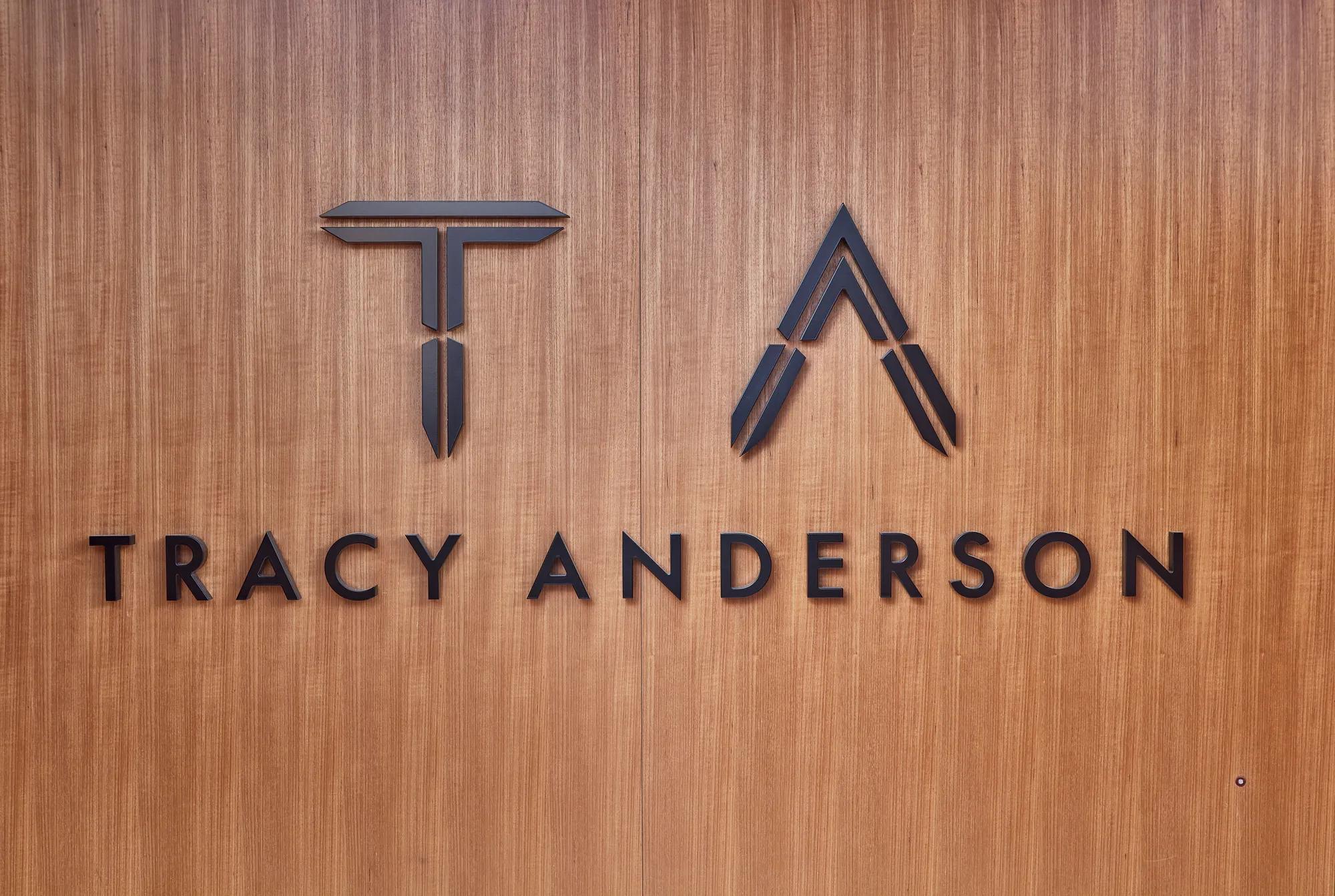 Tracy Anderson logo in black lettering and monogram on a warm wood-paneled wall.