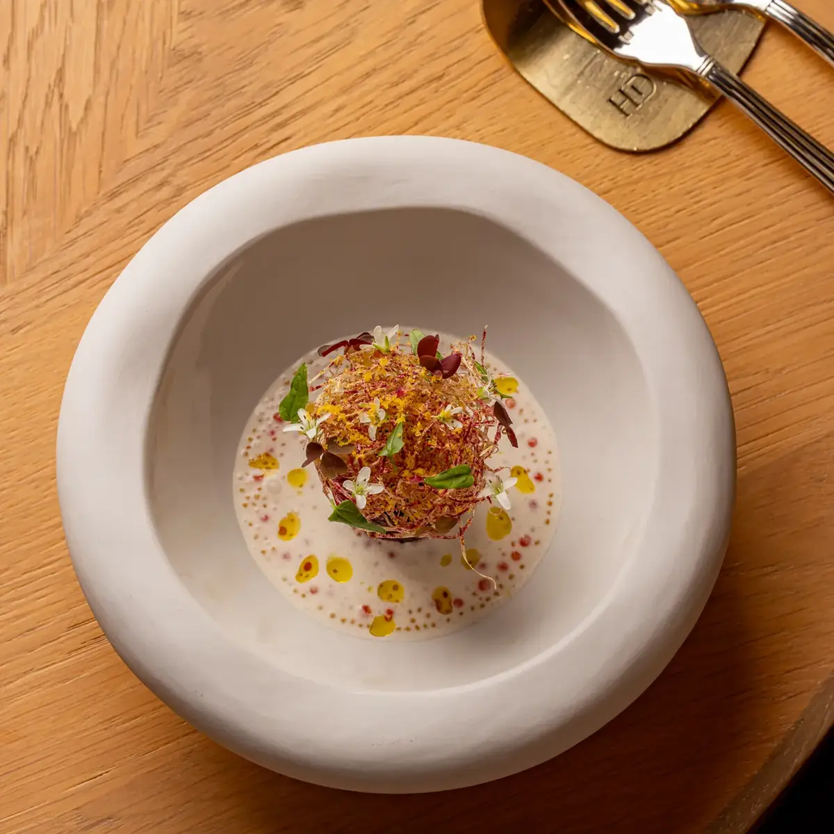 Gourmet plated dish with herbs and sauce in deep white bowl on wooden table beside cutlery.