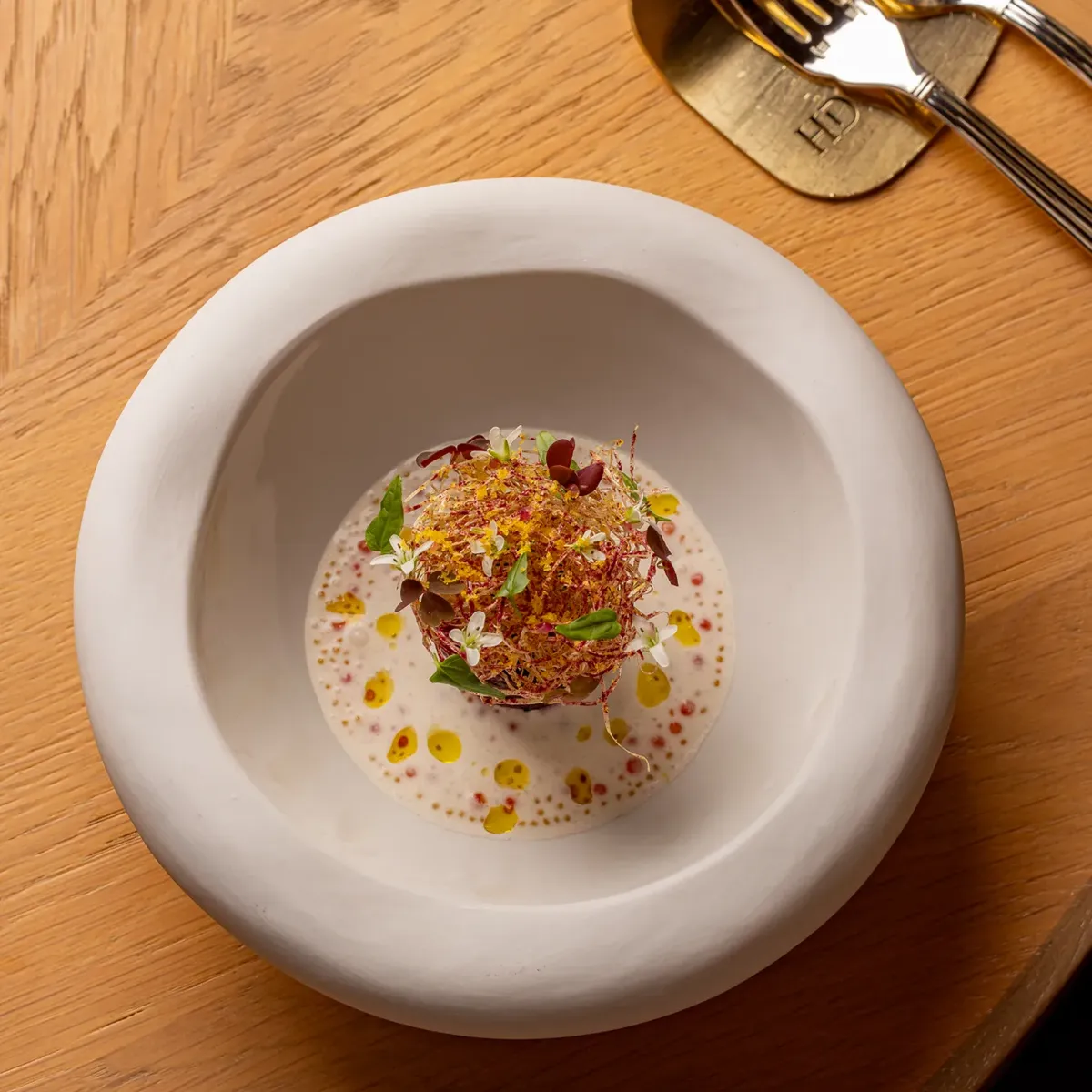 Gourmet plated dish with herbs and sauce in deep white bowl on wooden table beside cutlery.