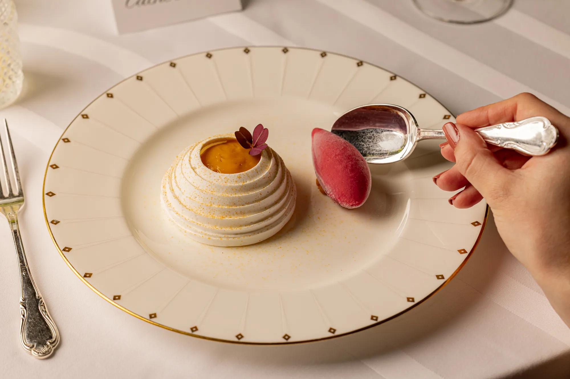 Elegant white meringue dessert with citrus centre, garnished with edible flowers, served on a fine china plate beside a raspberry sorbet quenelle and silver spoon.