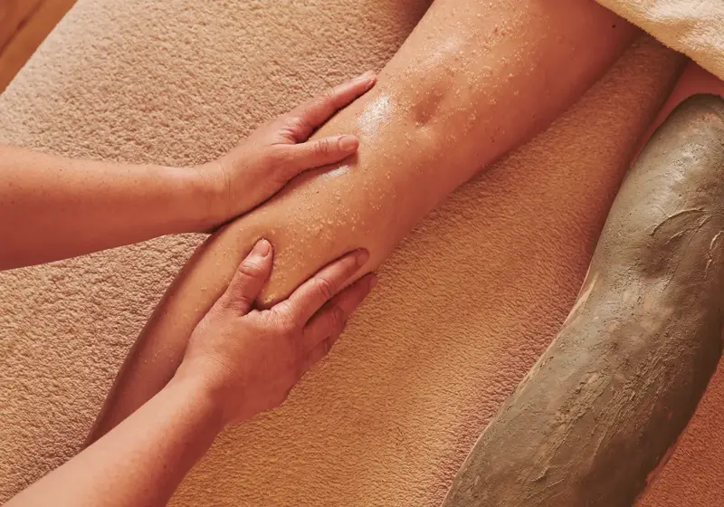 Person receiving a leg treatment, with one leg being massaged and the other covered in a green clay mask.