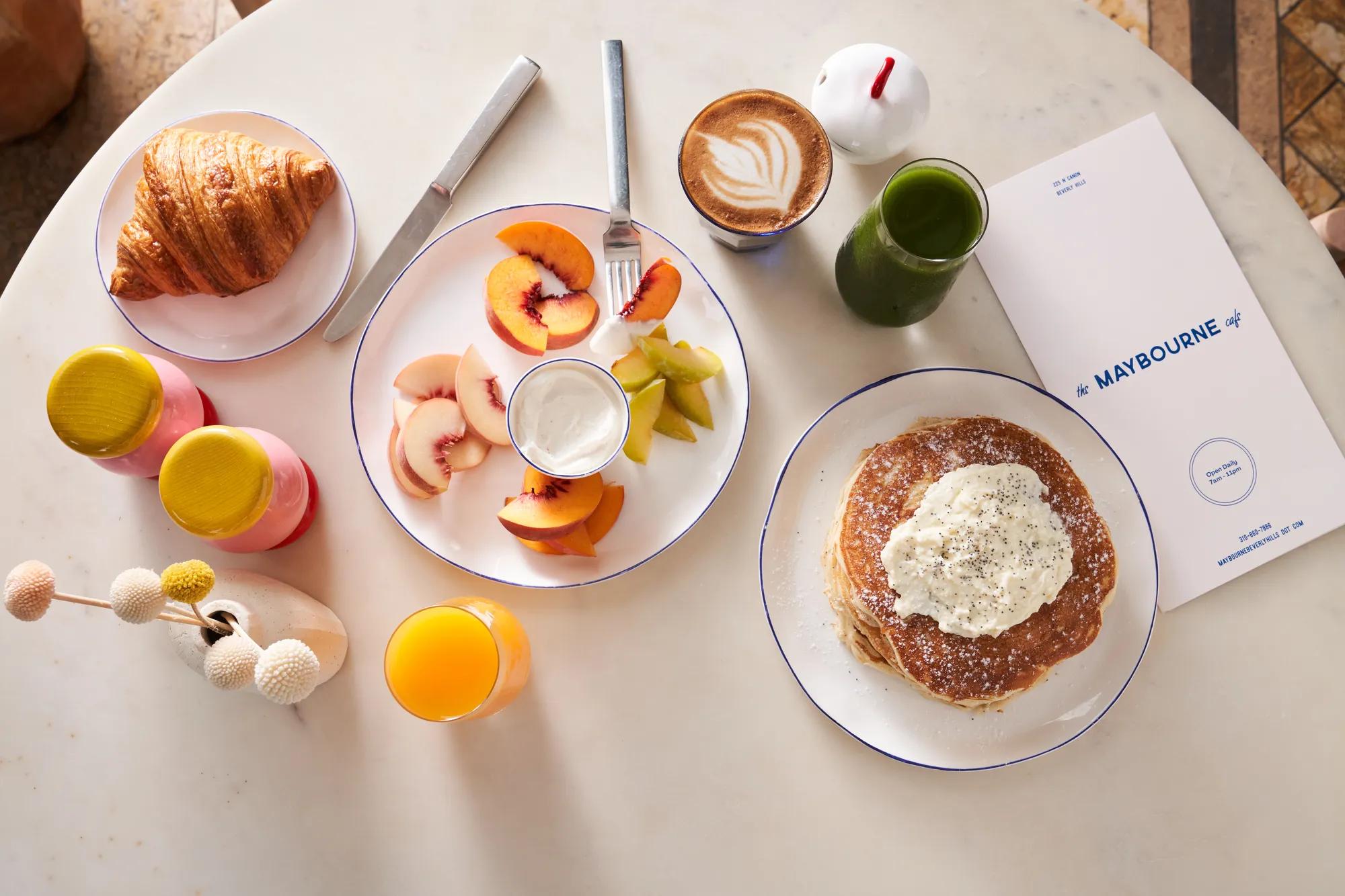 Overhead view of hotel breakfast with pancakes, fruit, croissant, coffee, juices, and a Maybourne-branded menu on a marble table.
