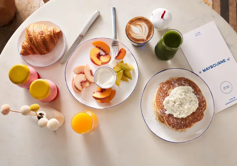 Overhead view of hotel breakfast with pancakes, fruit, croissant, coffee, juices, and a Maybourne-branded menu on a marble table.