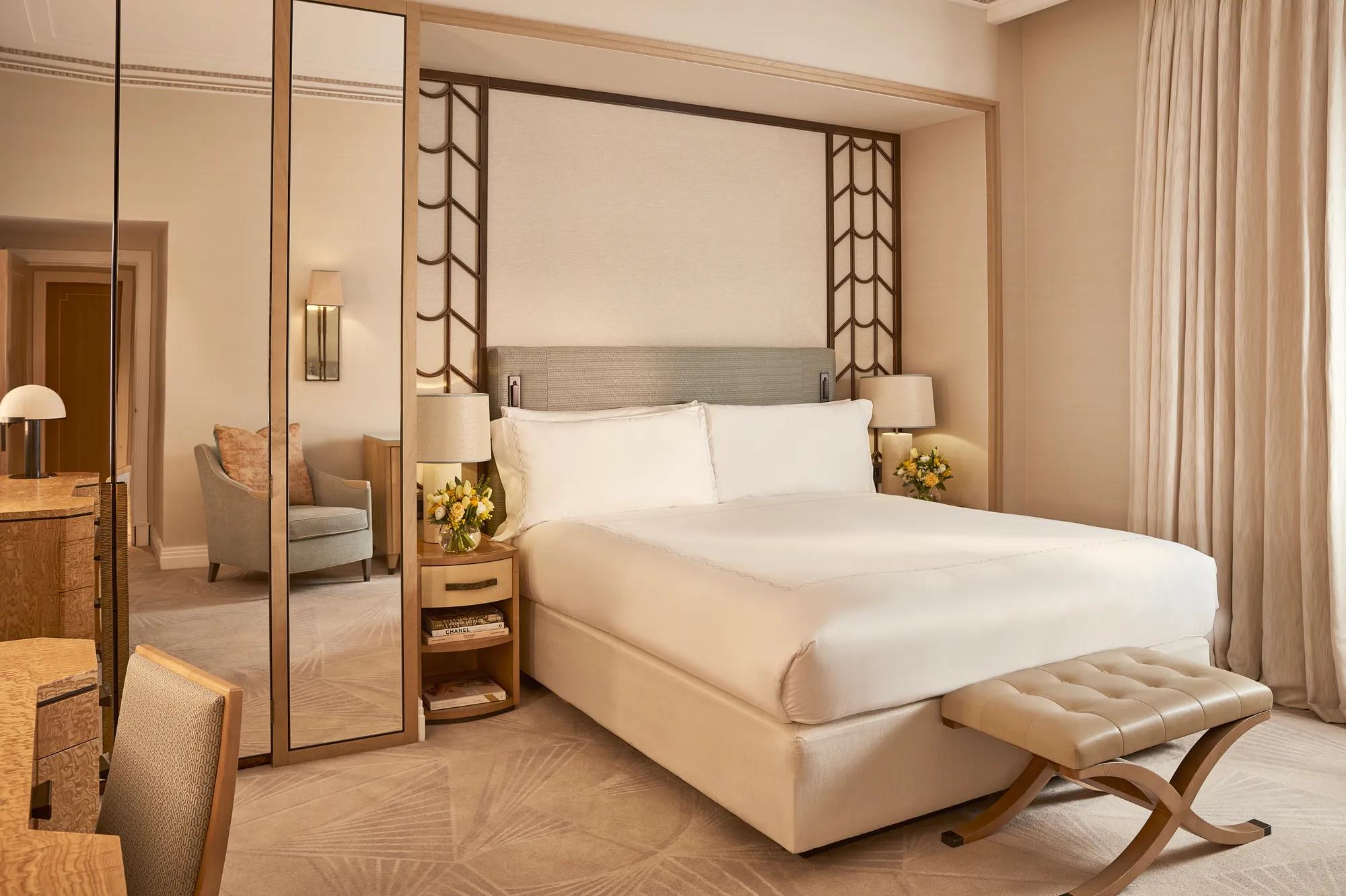 Elegant bedroom with a large bed dressed in white linens, mirrored panels, a padded bench at the foot, and light wood furnishings with soft neutral tones and natural light from a curtained window.