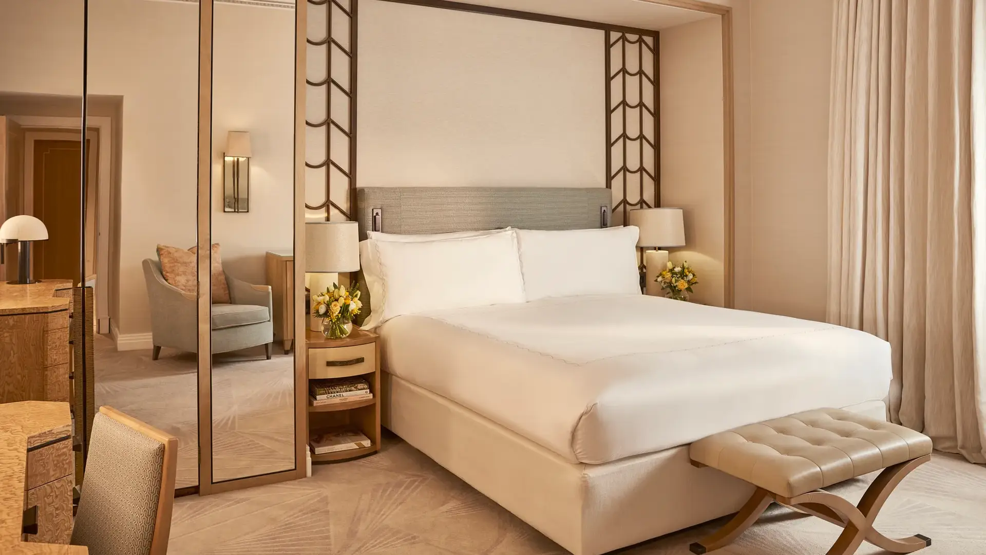 Elegant bedroom with a large bed dressed in white linens, mirrored panels, a padded bench at the foot, and light wood furnishings with soft neutral tones and natural light from a curtained window.