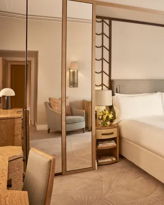 Elegant bedroom with a large bed dressed in white linens, mirrored panels, a padded bench at the foot, and light wood furnishings with soft neutral tones and natural light from a curtained window.