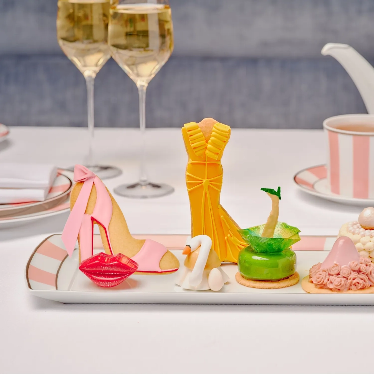 Pret A Portea Afternoon Tea platter of fashion-themed pastries—heel, gown, swan, green dress—with pink-striped china.