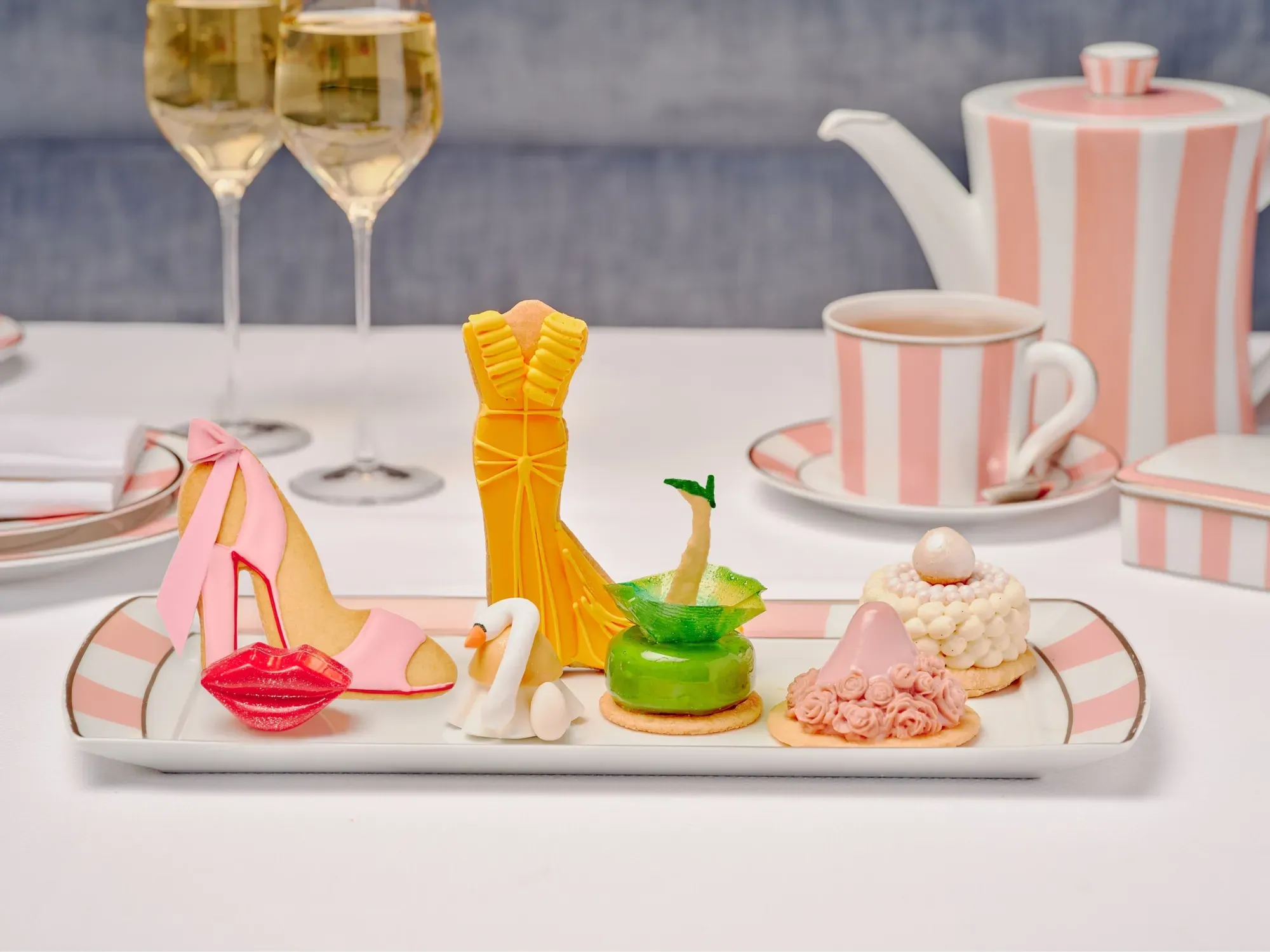Pret A Portea Afternoon Tea platter of fashion-themed pastries—heel, gown, swan, green dress—with pink-striped china.