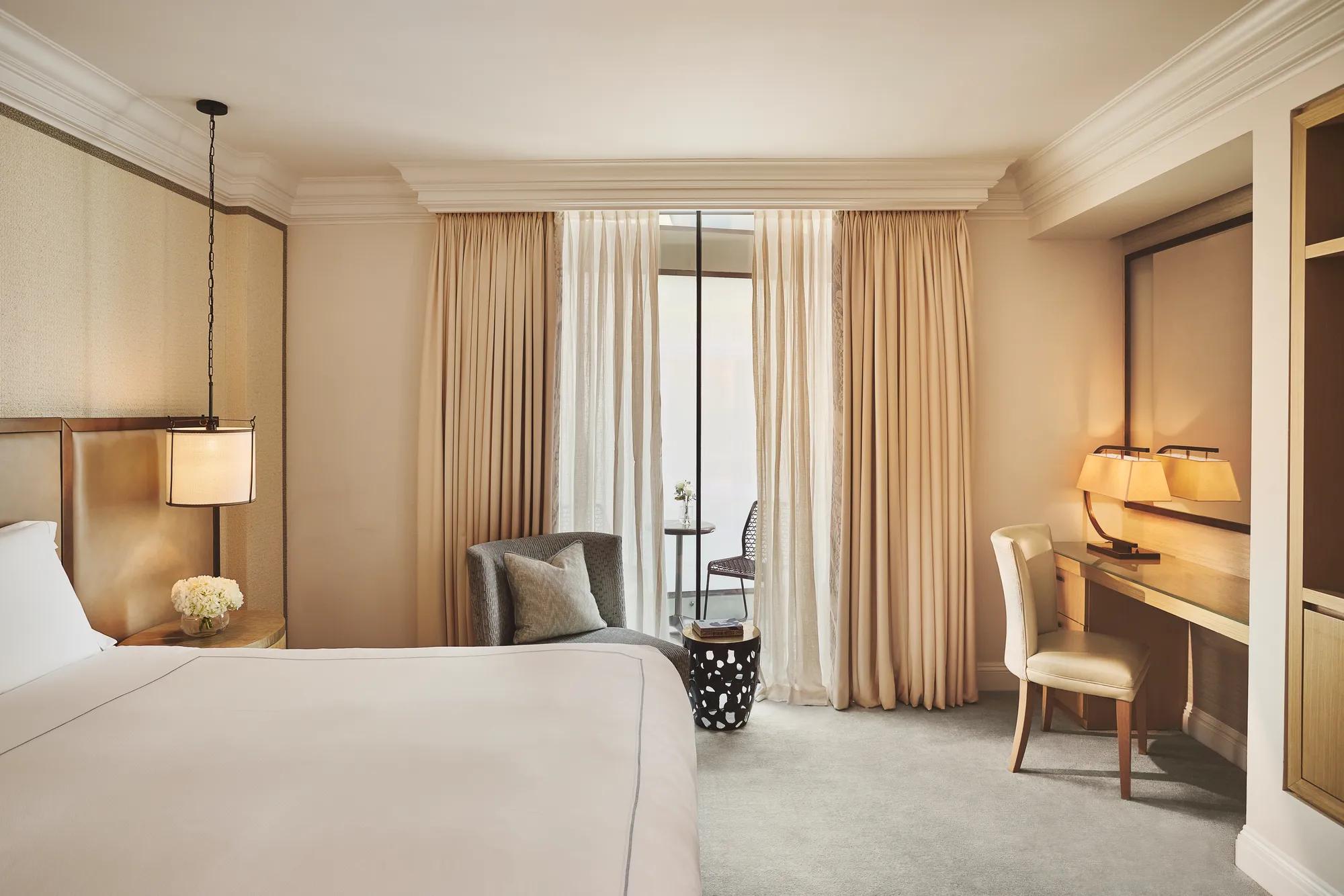 Bedroom with a neatly made bed, armchair, desk and chair beside floor-to-ceiling curtains leading to a balcony.
