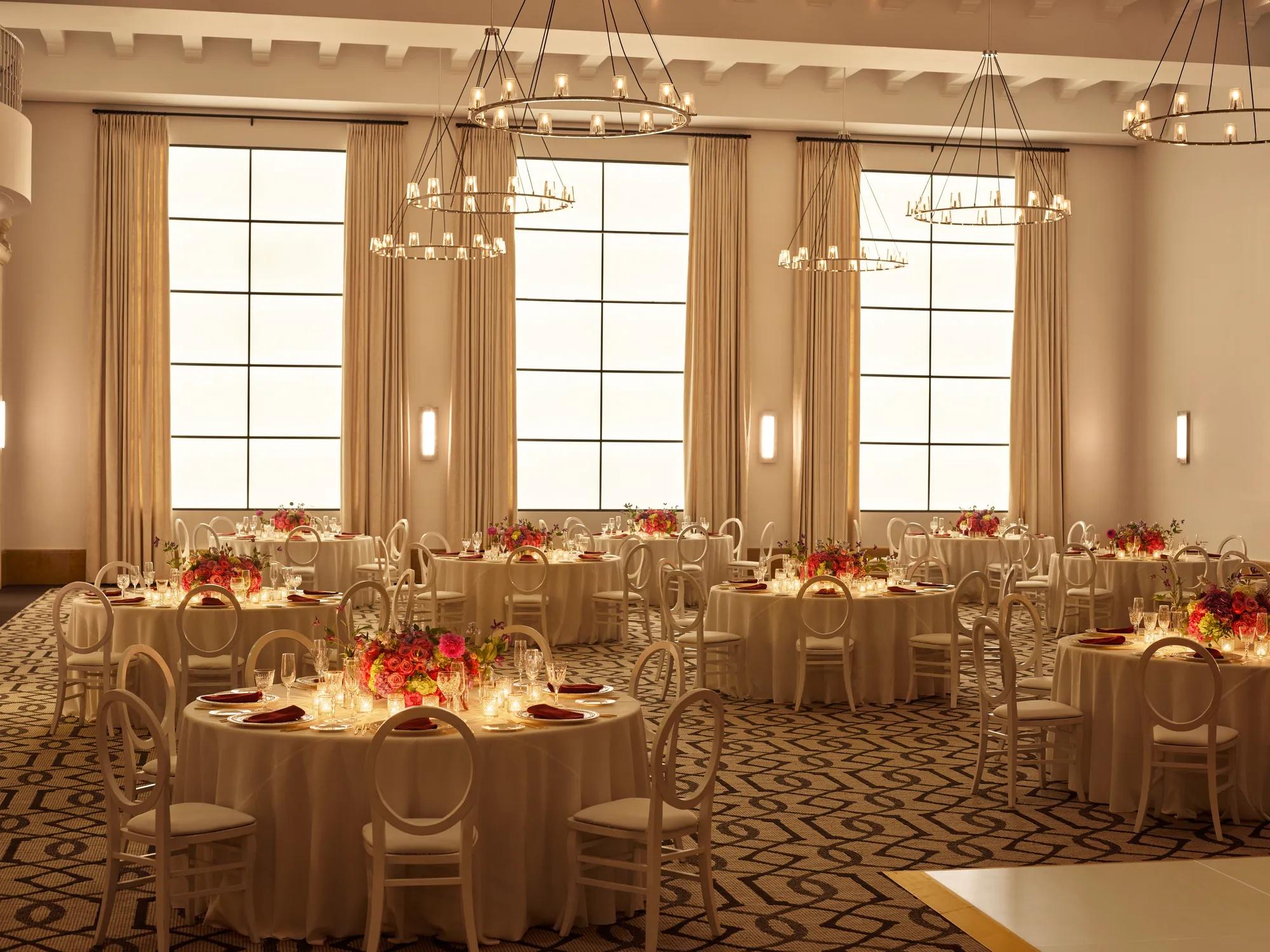 Beverly Hills Ballroom set for dinner with candlelit round tables, white chairs, and bright floral centerpieces beneath ring chandeliers.