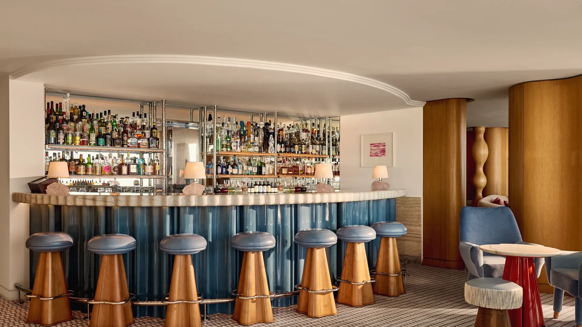 Bar élégant d’hôtel avec un comptoir incurvé habillé de panneaux bleus, des tabourets en bois aux assises en cuir bleu, et des étagères miroirs présentant une vaste collection de spiritueux.