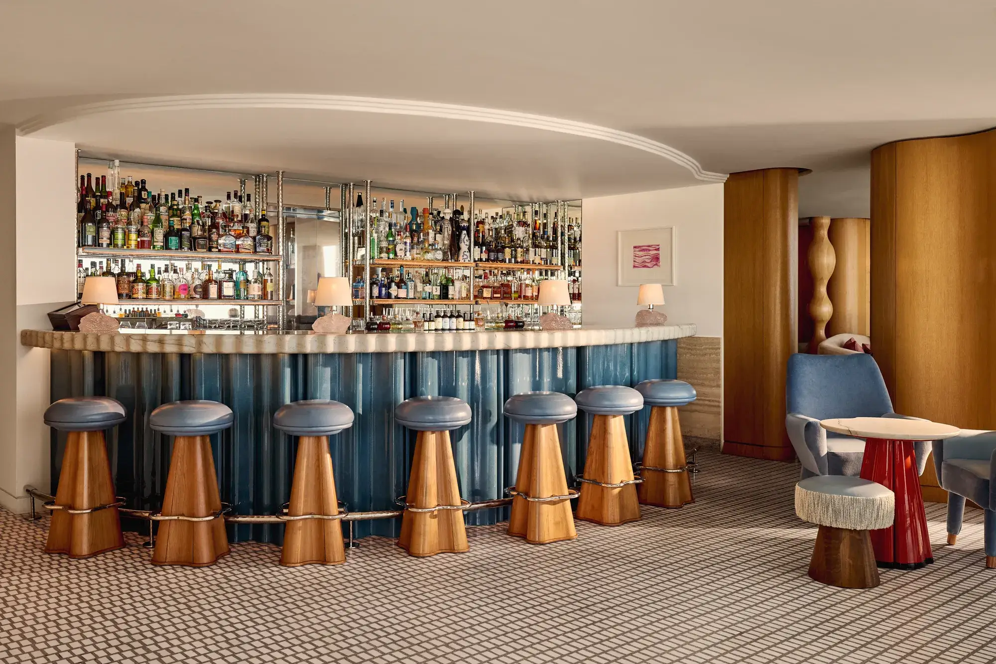 Elegant hotel bar with a curved counter clad in blue panels, wooden stools with blue leather seats, and mirrored shelves displaying an extensive collection of spirits.