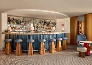 Bar élégant d’hôtel avec un comptoir incurvé habillé de panneaux bleus, des tabourets en bois aux assises en cuir bleu, et des étagères miroirs présentant une vaste collection de spiritueux.