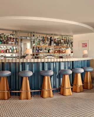 Elegant hotel bar with a curved counter clad in blue panels, wooden stools with blue leather seats, and mirrored shelves displaying an extensive collection of spirits.