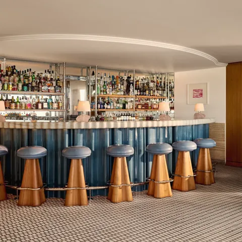 Elegant hotel bar with a curved counter clad in blue panels, wooden stools with blue leather seats, and mirrored shelves displaying an extensive collection of spirits.