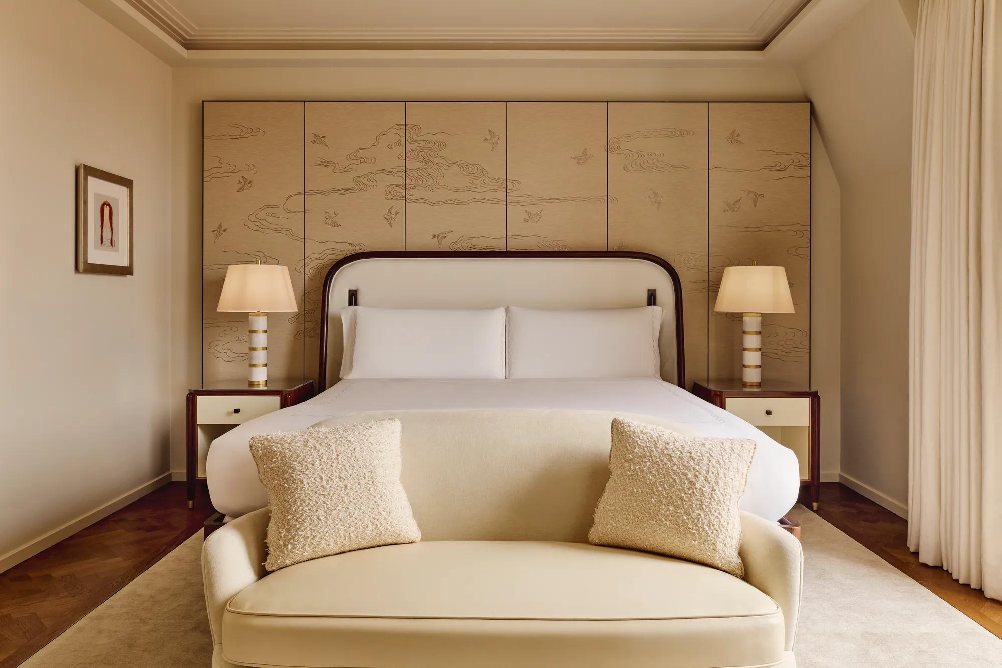 Serene bedroom with a king-size bed framed by a cream upholstered headboard, bedside tables with striped lamps, and a decorative wall panel featuring a subtle bird and cloud motif, with a soft cream loveseat at the foot of the bed.