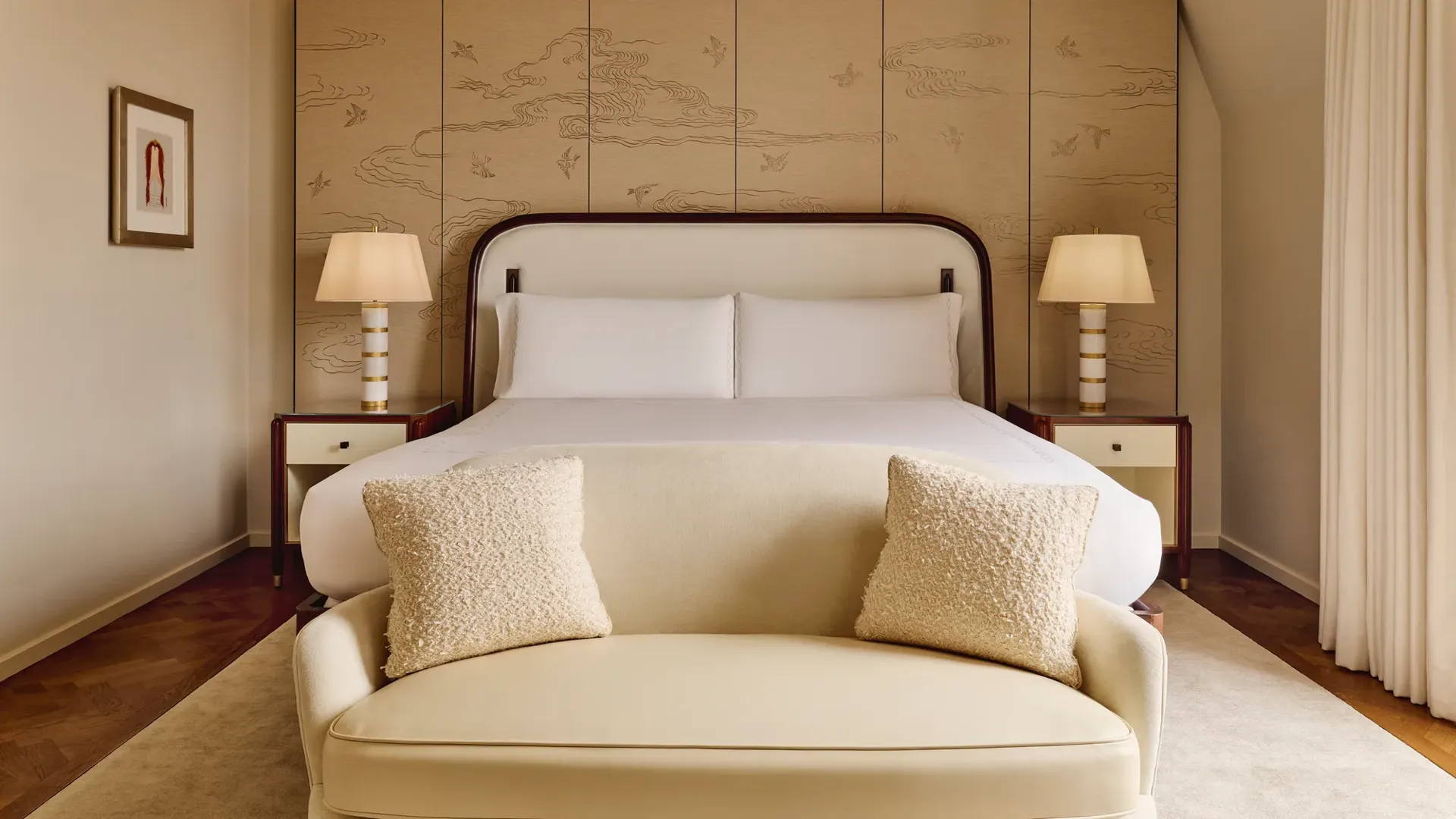 Serene bedroom with a king-size bed framed by a cream upholstered headboard, bedside tables with striped lamps, and a decorative wall panel featuring a subtle bird and cloud motif, with a soft cream loveseat at the foot of the bed.