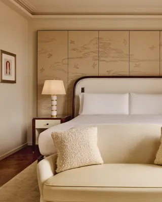 Serene bedroom with a king-size bed framed by a cream upholstered headboard, bedside tables with striped lamps, and a decorative wall panel featuring a subtle bird and cloud motif, with a soft cream loveseat at the foot of the bed.
