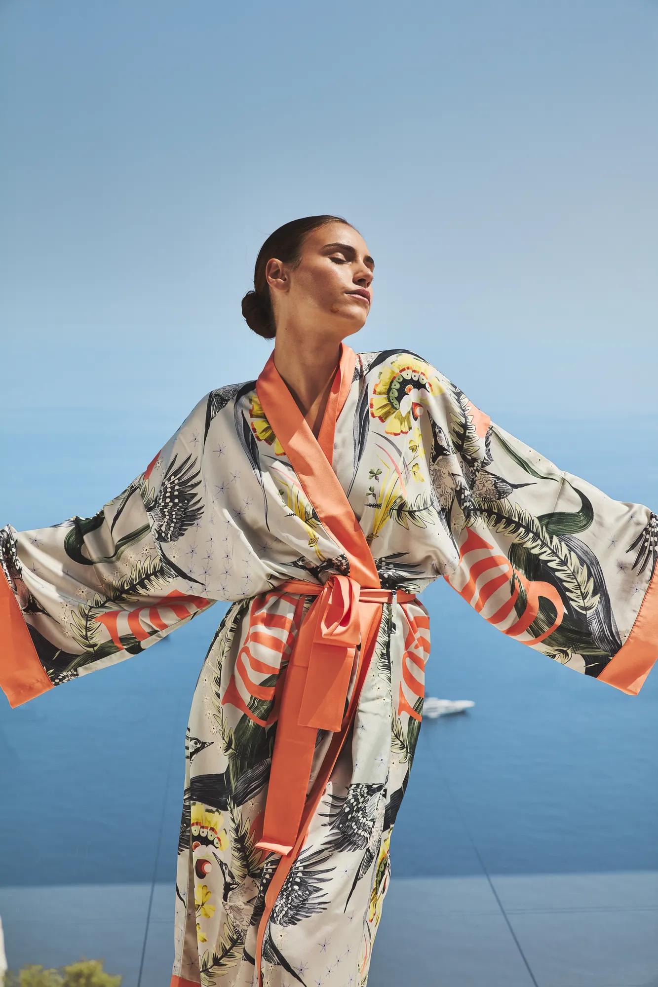 Woman wearing patterned robe with open arms, standing against clear blue sky and Mediterranean sea backdrop.
