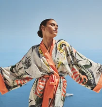 Woman wearing patterned robe with open arms, standing against clear blue sky and Mediterranean sea backdrop.