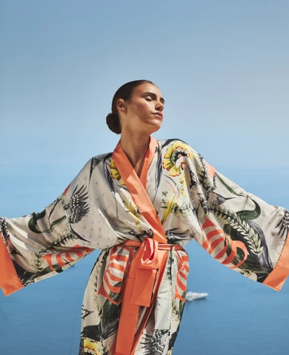 Woman wearing patterned robe with open arms, standing against clear blue sky and Mediterranean sea backdrop.