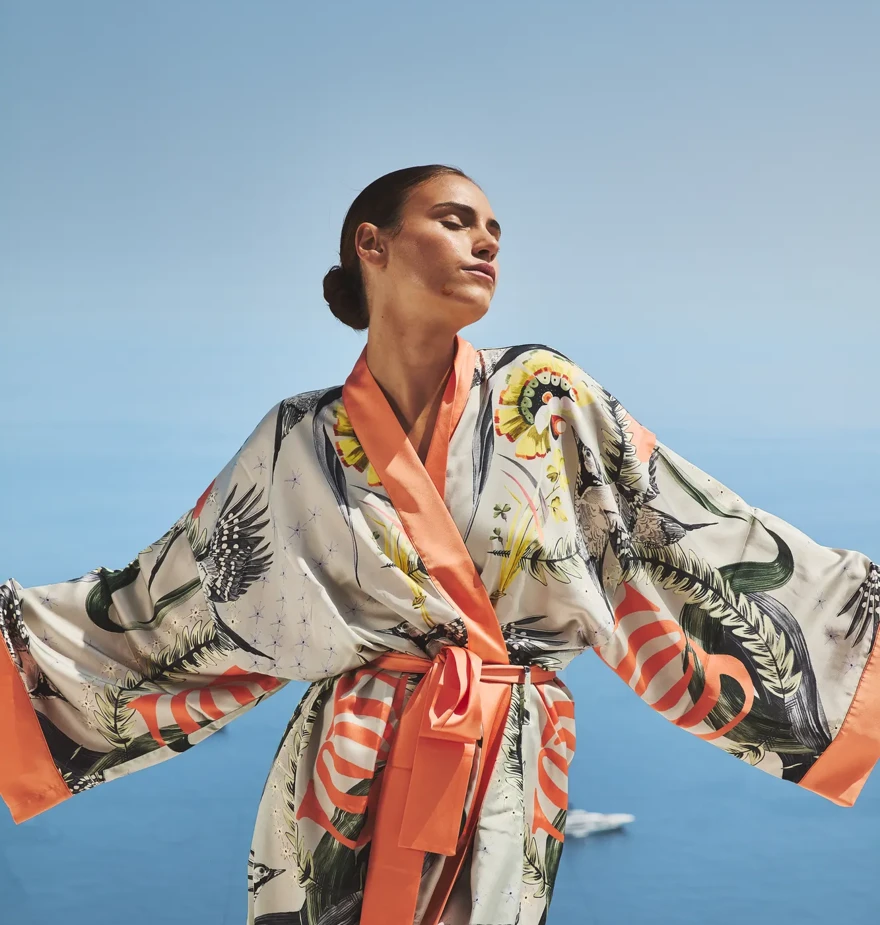 Woman wearing patterned robe with open arms, standing against clear blue sky and Mediterranean sea backdrop.