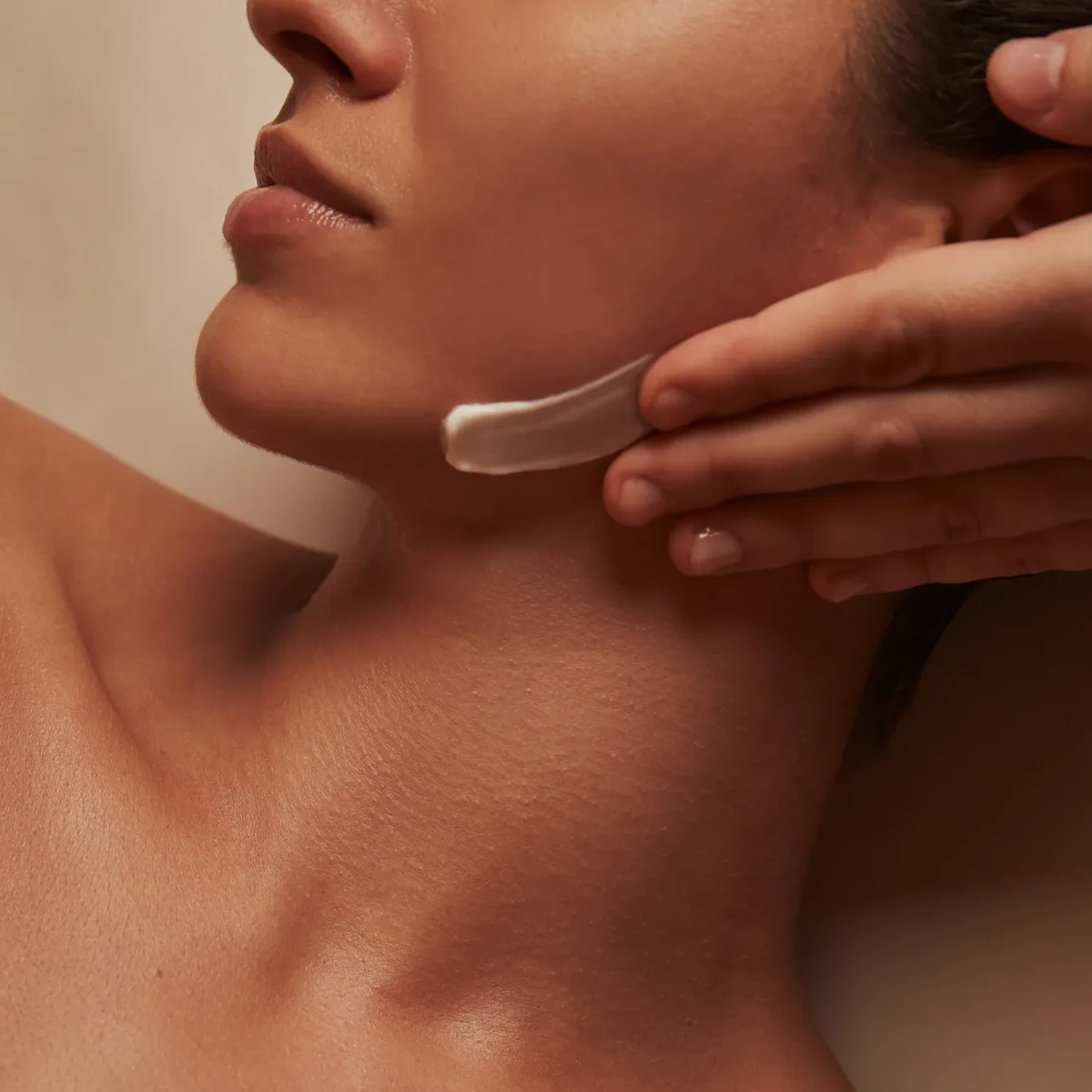 Close-up of therapist applying cream to woman’s jawline during facial treatment, warm lighting highlighting smooth skin and relaxed expression.