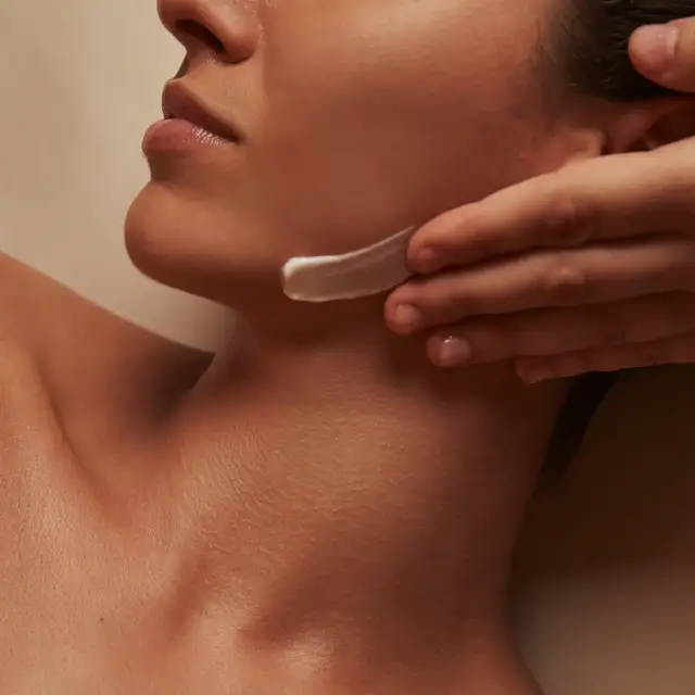 Close-up of therapist applying cream to woman’s jawline during facial treatment, warm lighting highlighting smooth skin and relaxed expression.