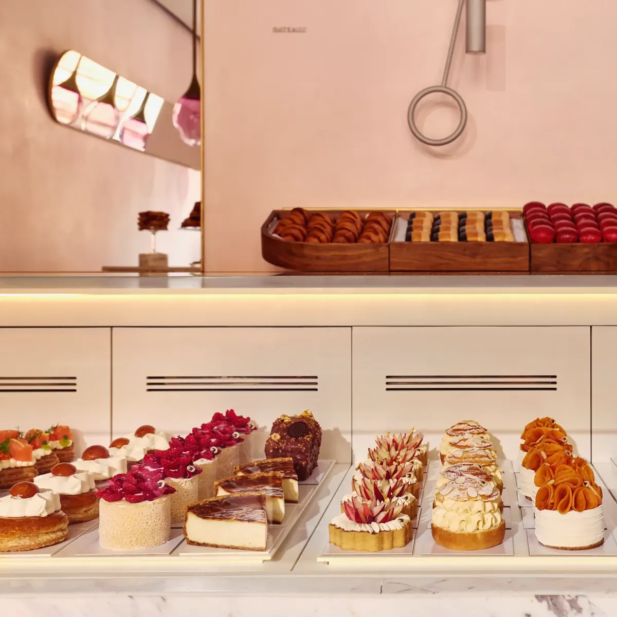 Display case of assorted pastries and tarts in a pastel-pink patisserie, with rows of colorful, finely decorated desserts.