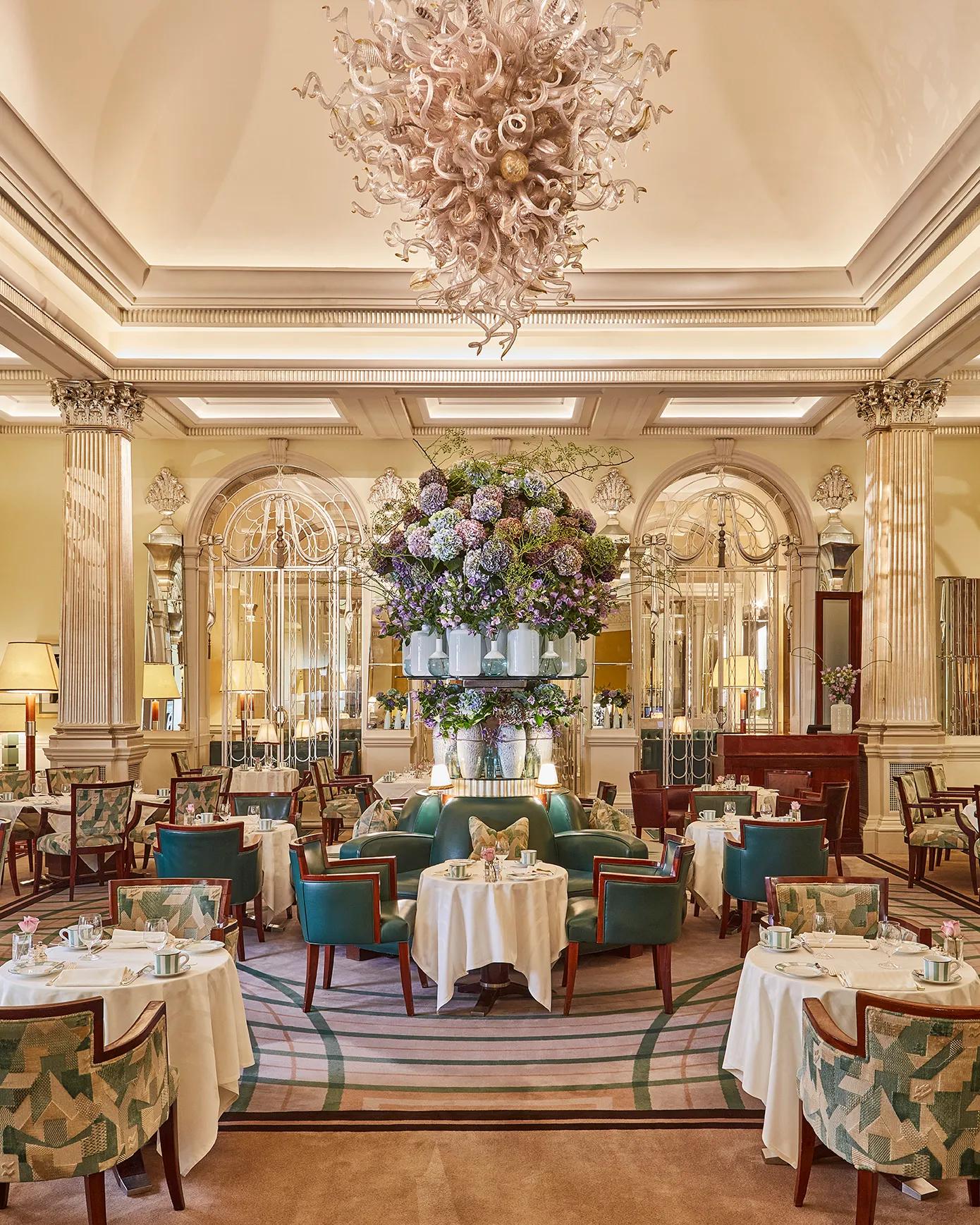 Elegant dining room at Claridge’s with round tables, teal chairs and a large floral display beneath a sculptural chandelier.