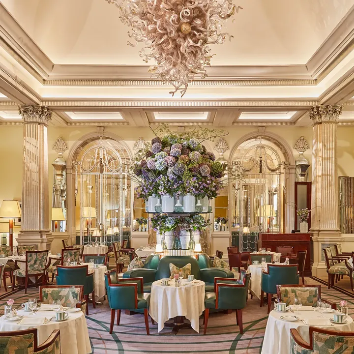 Elegant dining room at Claridge’s with round tables, teal chairs and a large floral display beneath a sculptural chandelier.