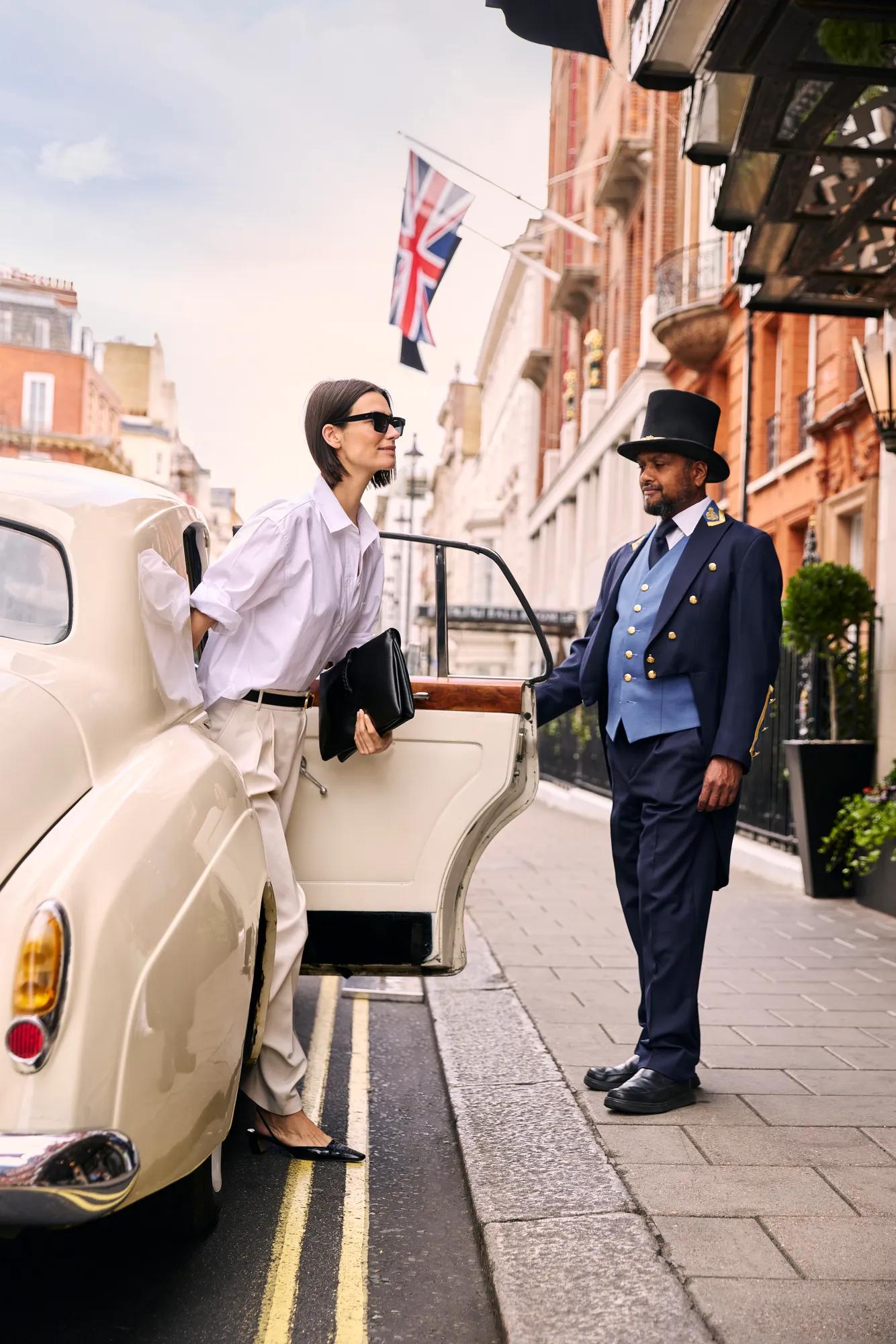 Guest stepping from a classic cream-coloured car outside a grand London hotel, welcomed on a flag-lined street by a uniformed doorman.
