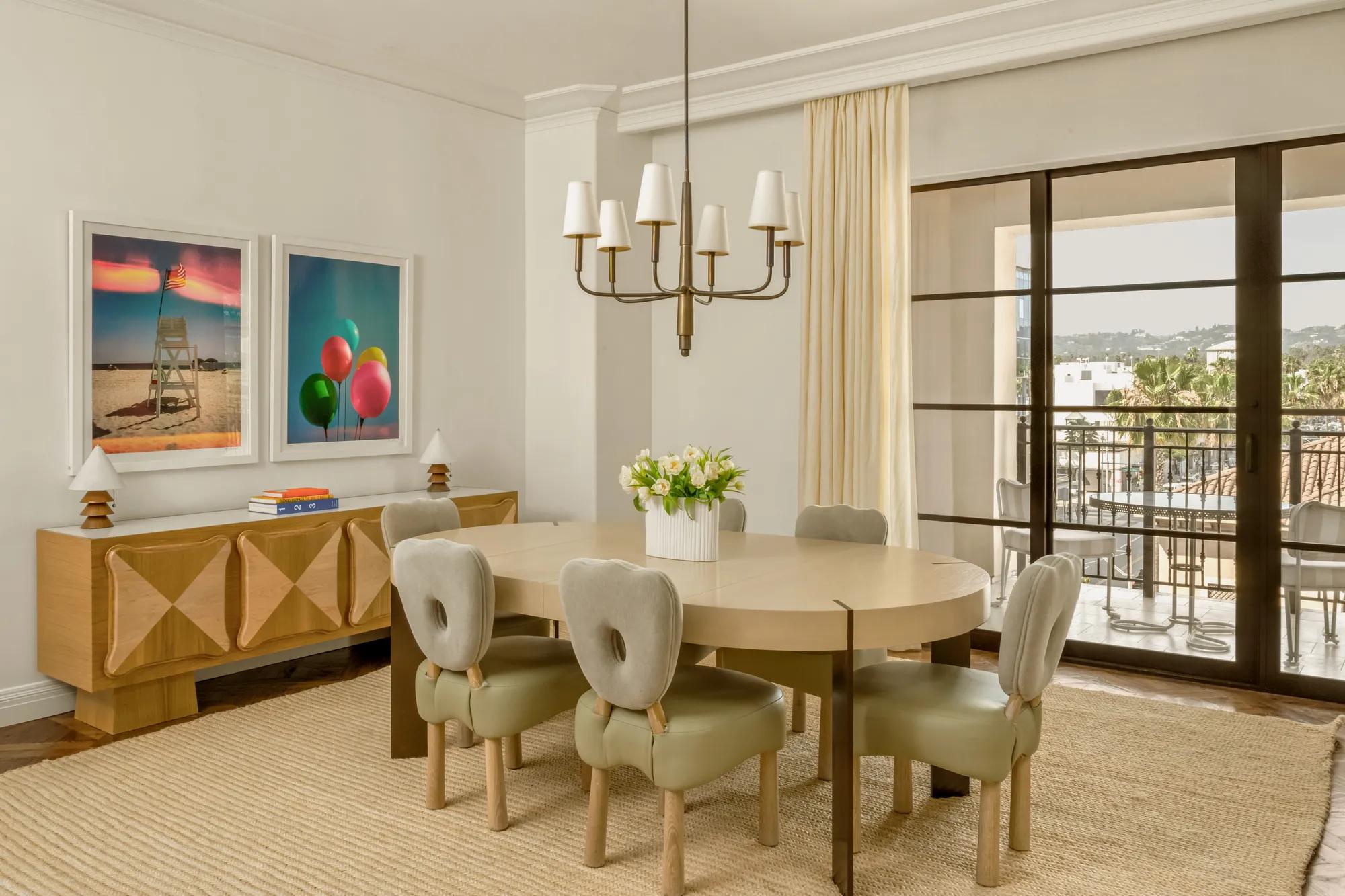 Elegant dining room with oval table, upholstered chairs, modern artwork, chandelier, and balcony views through large glass doors.