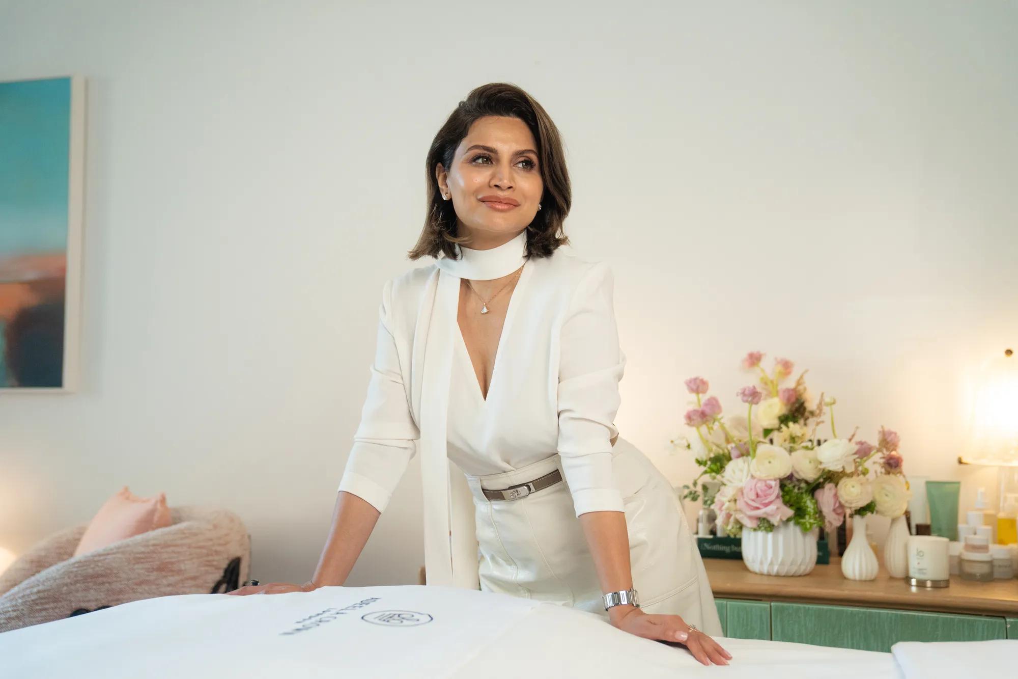 Person in white outfit leaning over a spa table in a softly lit room with flowers, skincare products, and minimalist decor.