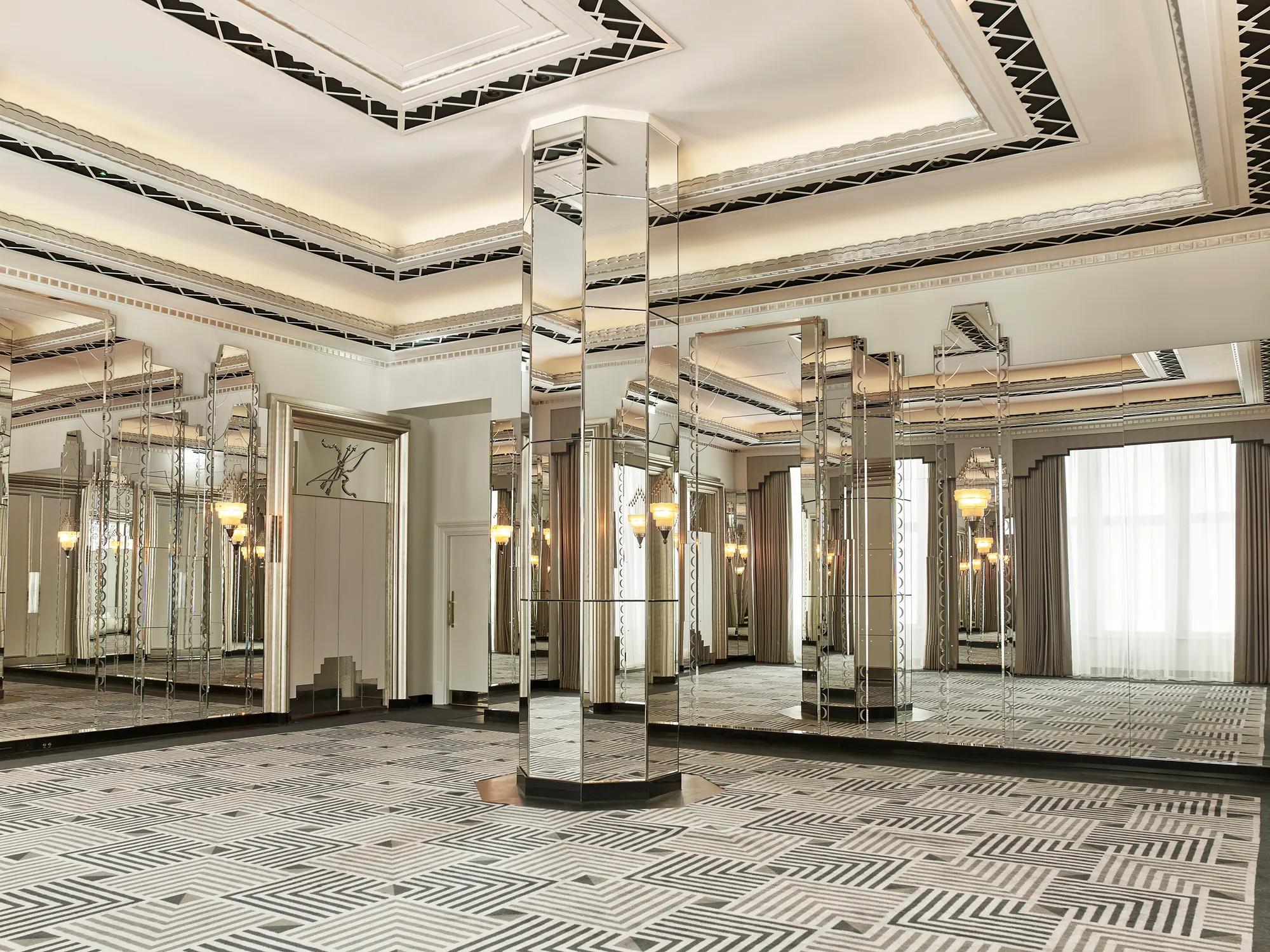 An elegant Art Deco–style ballroom featuring mirrored columns, geometric patterned carpet, and decorative ceiling details, with large windows framed by floor-length curtains allowing natural light to fill the space.