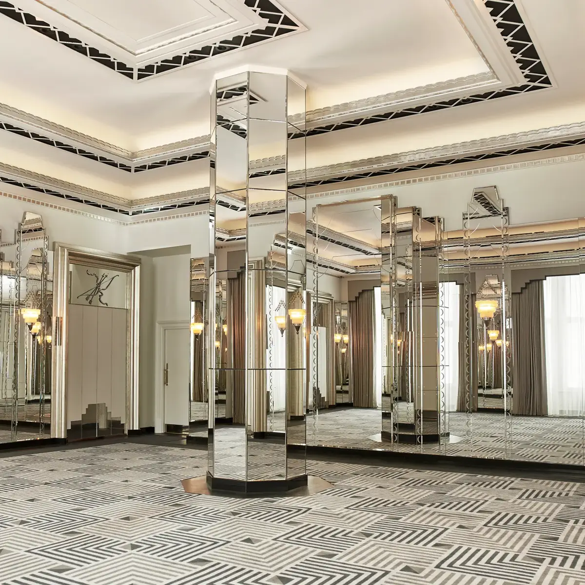 An elegant Art Deco–style ballroom featuring mirrored columns, geometric patterned carpet, and decorative ceiling details, with large windows framed by floor-length curtains allowing natural light to fill the space.