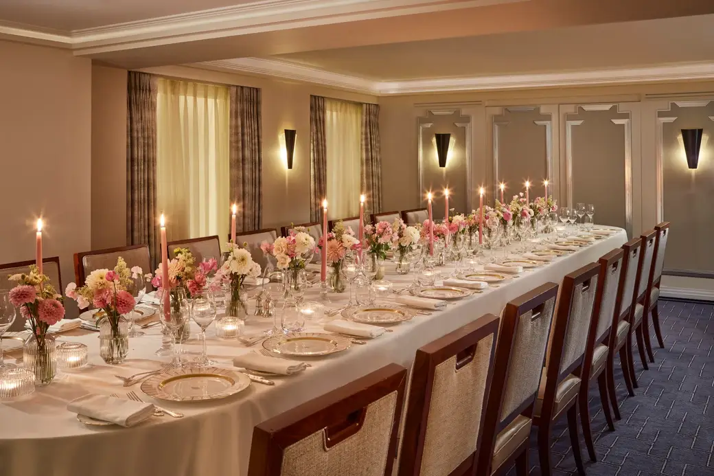 Elegant private dining setup at Claridge’s with a long candlelit table surrounded by upholstered chairs, decorated with pink and white floral arrangements and fine china on crisp white linen.