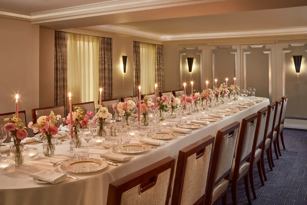 Elegant private dining setup at Claridge’s with a long candlelit table surrounded by upholstered chairs, decorated with pink and white floral arrangements and fine china on crisp white linen.