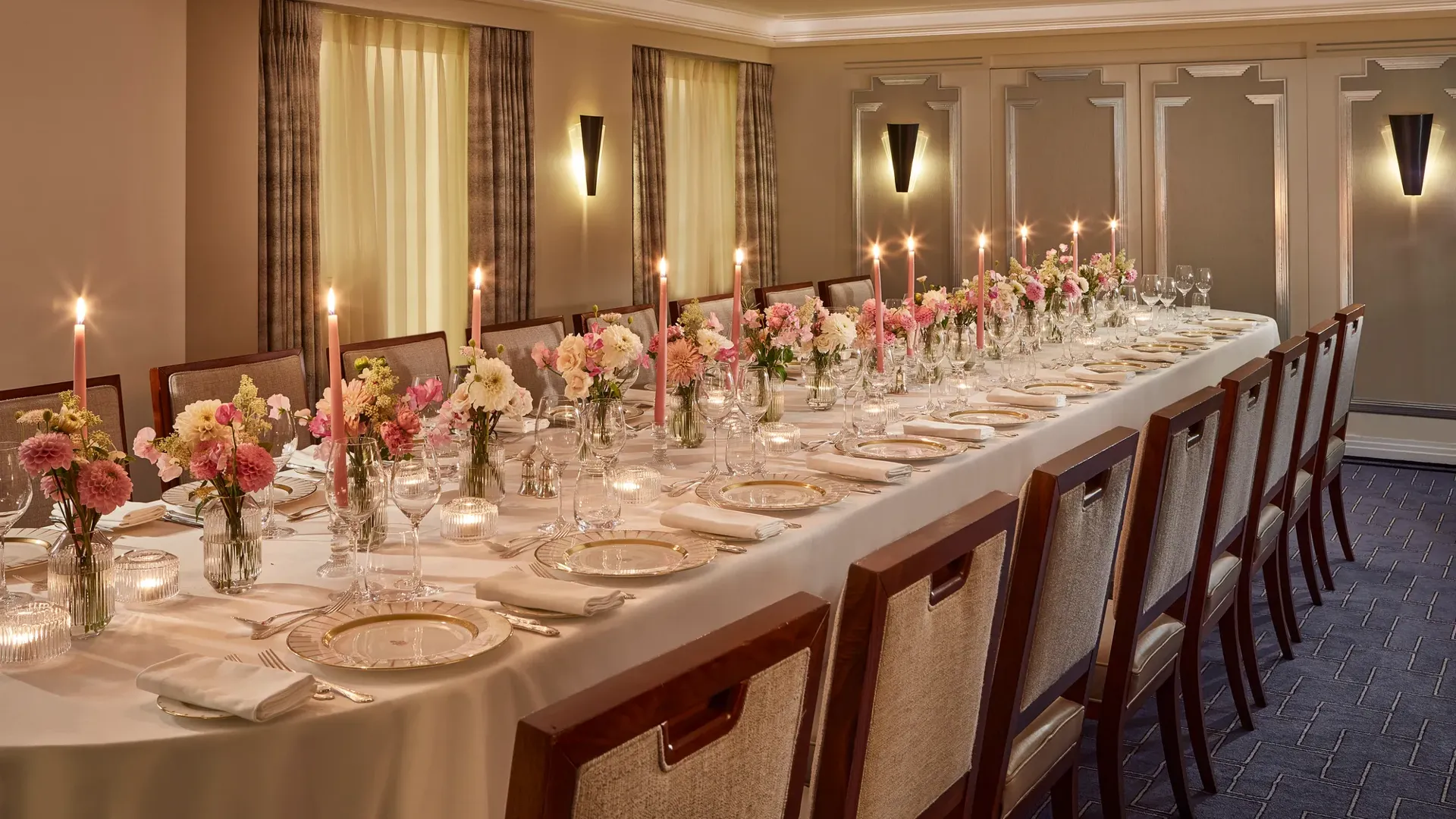 Elegant private dining setup at Claridge’s with a long candlelit table surrounded by upholstered chairs, decorated with pink and white floral arrangements and fine china on crisp white linen.