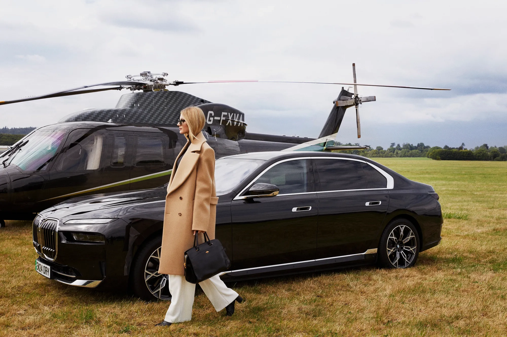 Woman walks away from black helicopter and luxury car across grassy airfield, dressed in white trousers and camel coat.
