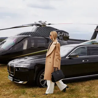 Woman walks away from black helicopter and luxury car across grassy airfield, dressed in white trousers and camel coat.