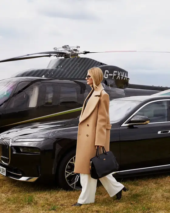 Woman walks away from black helicopter and luxury car across grassy airfield, dressed in white trousers and camel coat.