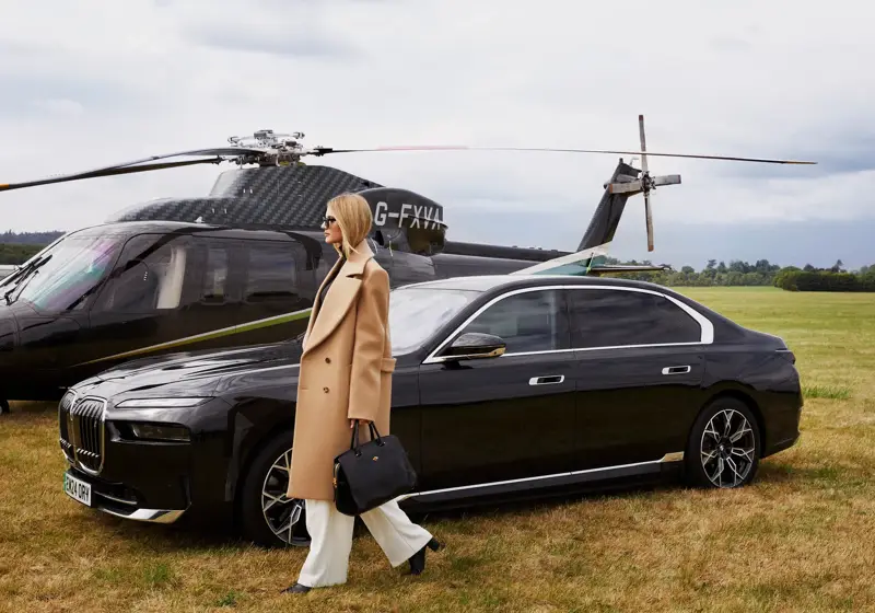 Woman walks away from black helicopter and luxury car across grassy airfield, dressed in white trousers and camel coat.