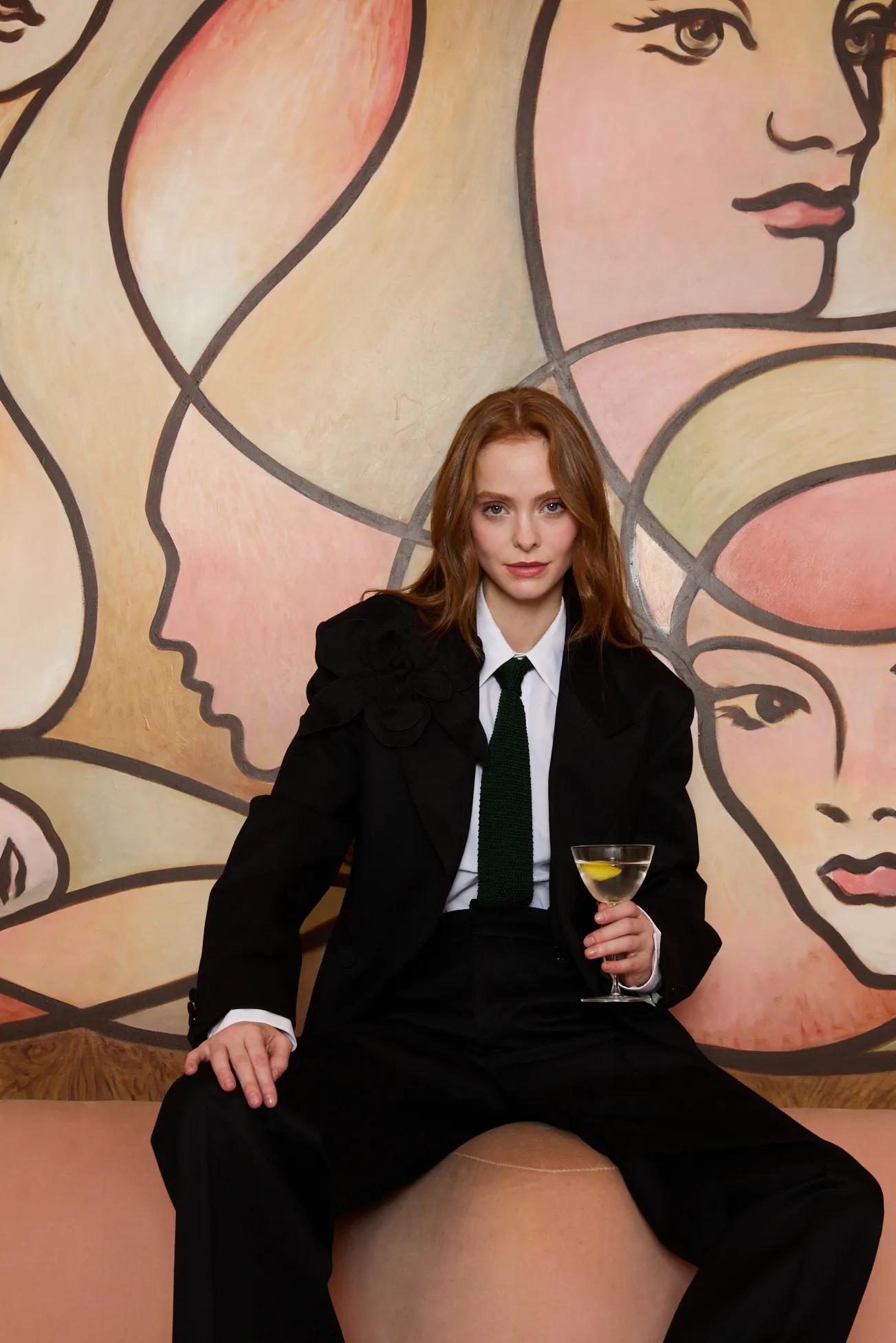 A person in an oversized black suit, white shirt, and dark green tie sits on a curved blush-colored bench, holding a stemmed cocktail glass.