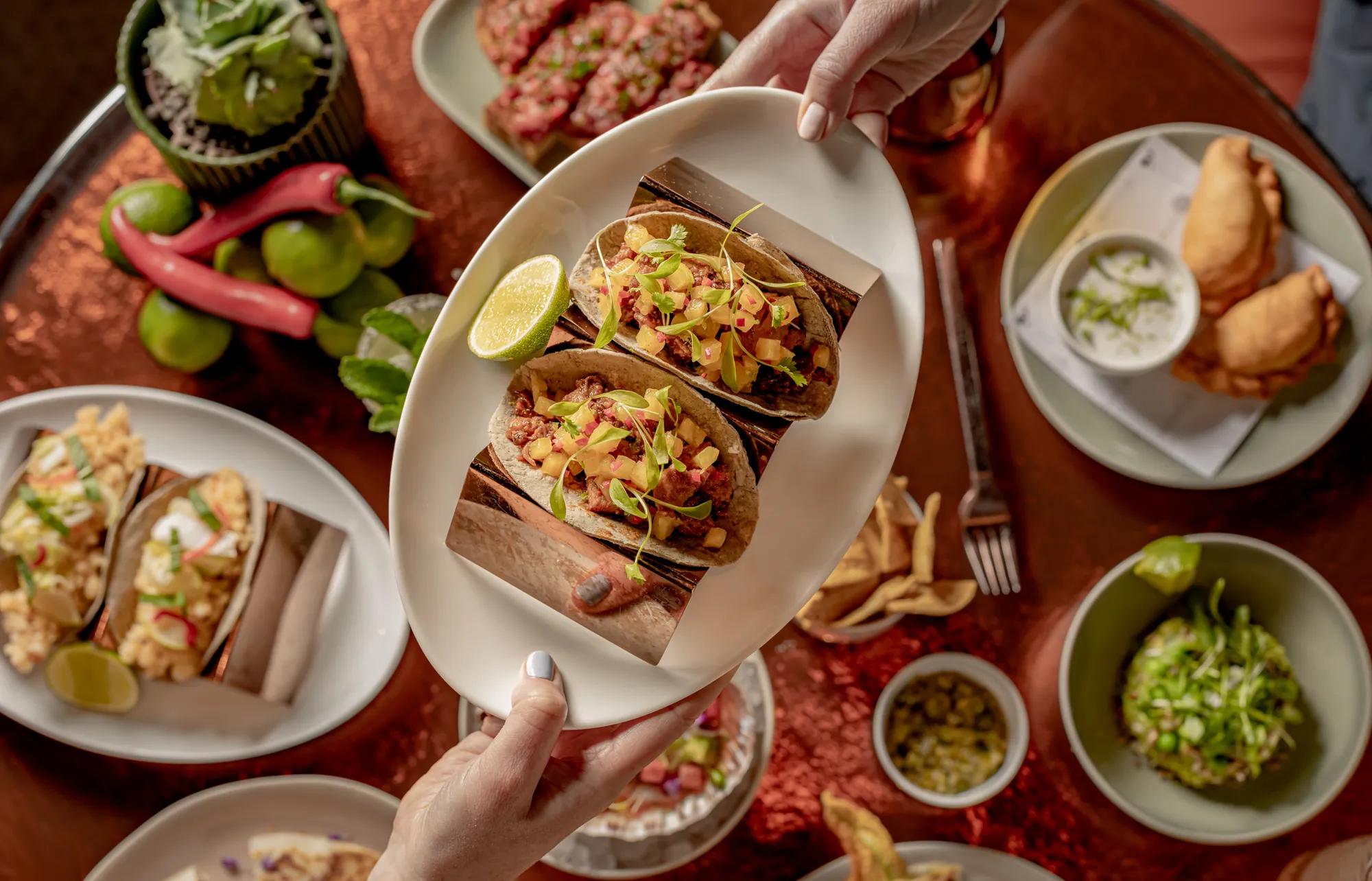 Overhead view of tacos garnished with fresh herbs, surrounded by small plates of appetisers, dips, vegetables and lime on a red tablecloth.