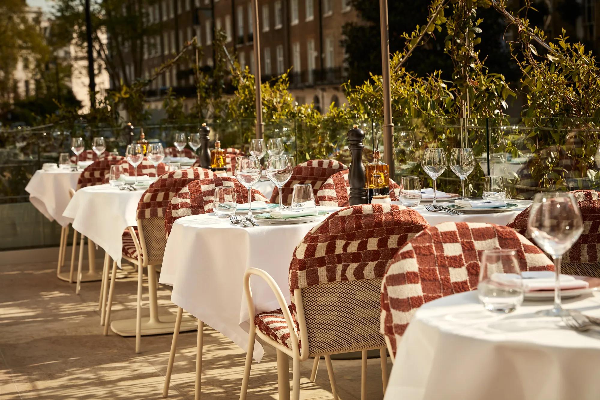 Alfresco dining setup with white tablecloths, glassware, and patterned chairs overlooking a green, leafy Mayfair street view