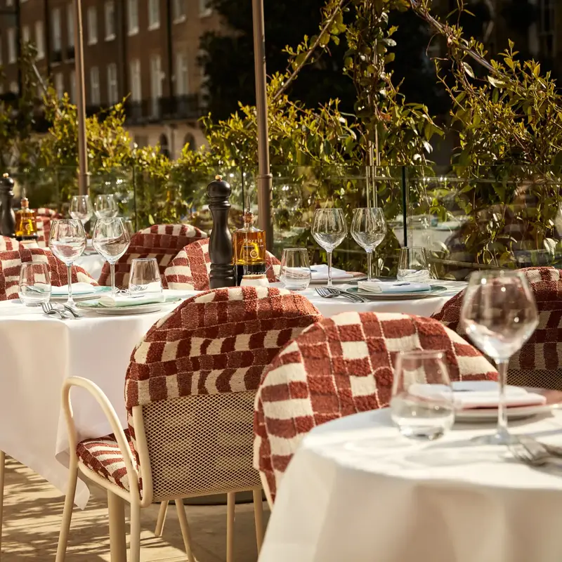Alfresco dining setup with white tablecloths, glassware, and patterned chairs overlooking a green, leafy Mayfair street view