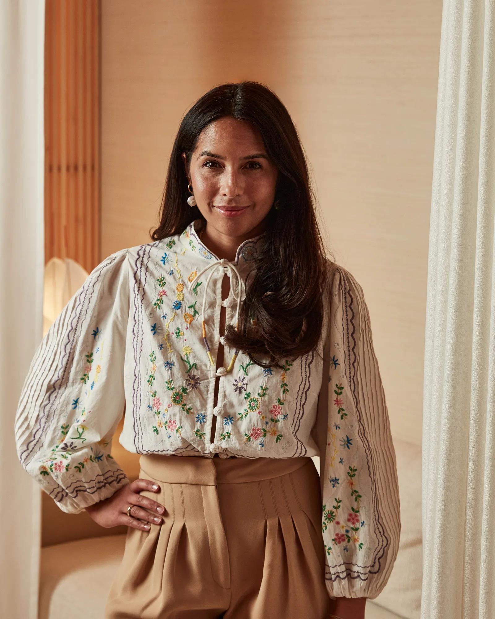 Woman wearing embroidered blouse and high-waisted trousers, standing indoors with hand on hip in softly lit neutral setting.