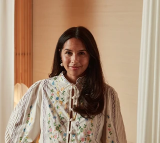 Woman wearing embroidered blouse and high-waisted trousers, standing indoors with hand on hip in softly lit neutral setting.