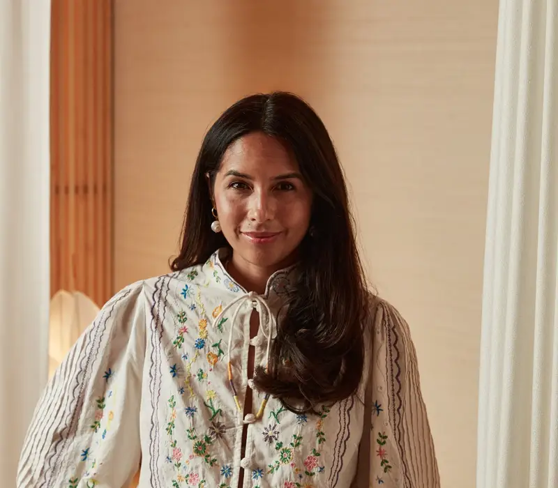 Woman wearing embroidered blouse and high-waisted trousers, standing indoors with hand on hip in softly lit neutral setting.